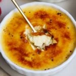 A close-up of creme brulee in a white ramekin, with a spoon breaking through the golden caramelized sugar crust to reveal the creamy custard inside