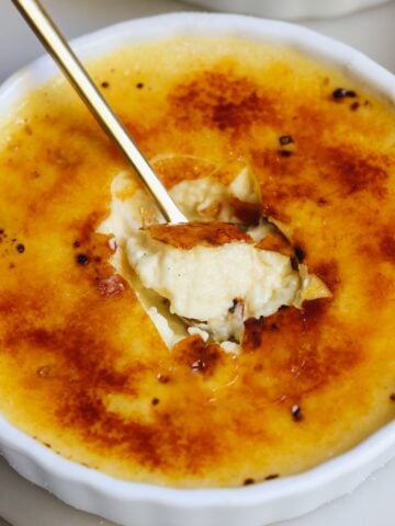 A close-up of creme brulee in a white ramekin, with a spoon breaking through the golden caramelized sugar crust to reveal the creamy custard inside