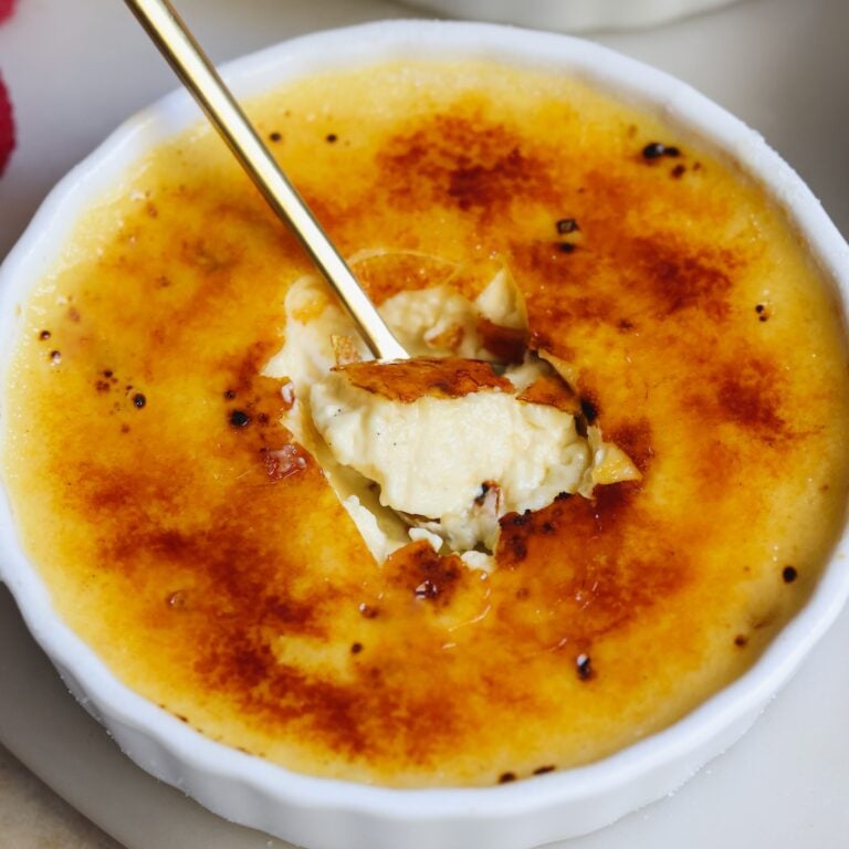 A close-up of creme brulee in a white ramekin, with a spoon breaking through the golden caramelized sugar crust to reveal the creamy custard inside