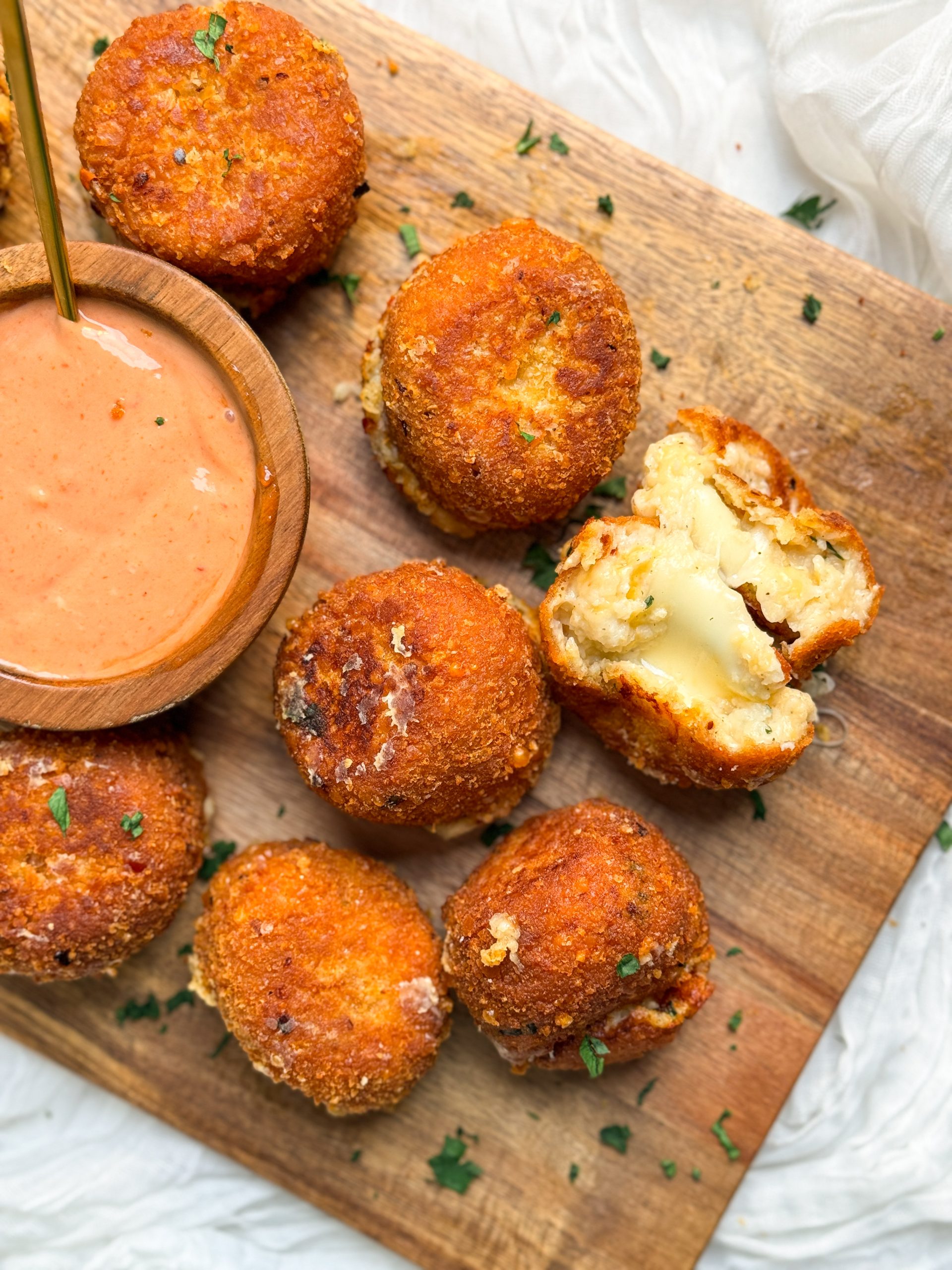crispy fried mashed potato balls on a cutting board, one ball is broken in half to show soft cheesy middle. balls are golden brown and crispy. small bowl with sauce on the side
