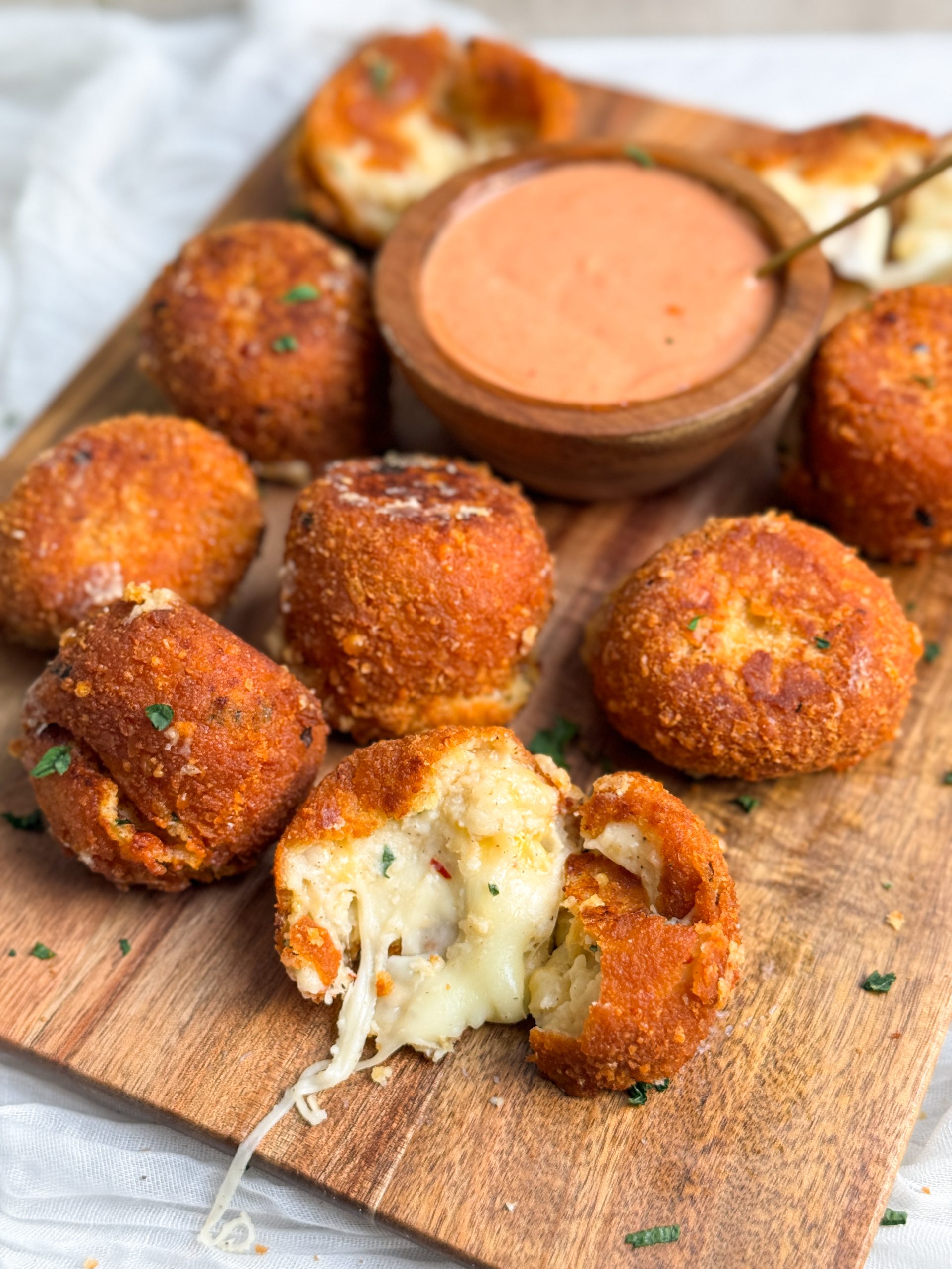 crispy fried mashed potato balls on a cutting board, one ball is broken in half to show soft cheesy middle. balls are golden brown and crispy. small bowl with sauce on the side. picture from an angle