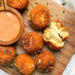 crispy fried mashed potato balls on a cutting board, one ball is broken in half to show soft cheesy middle. balls are golden brown and crispy. small bowl with sauce on the side