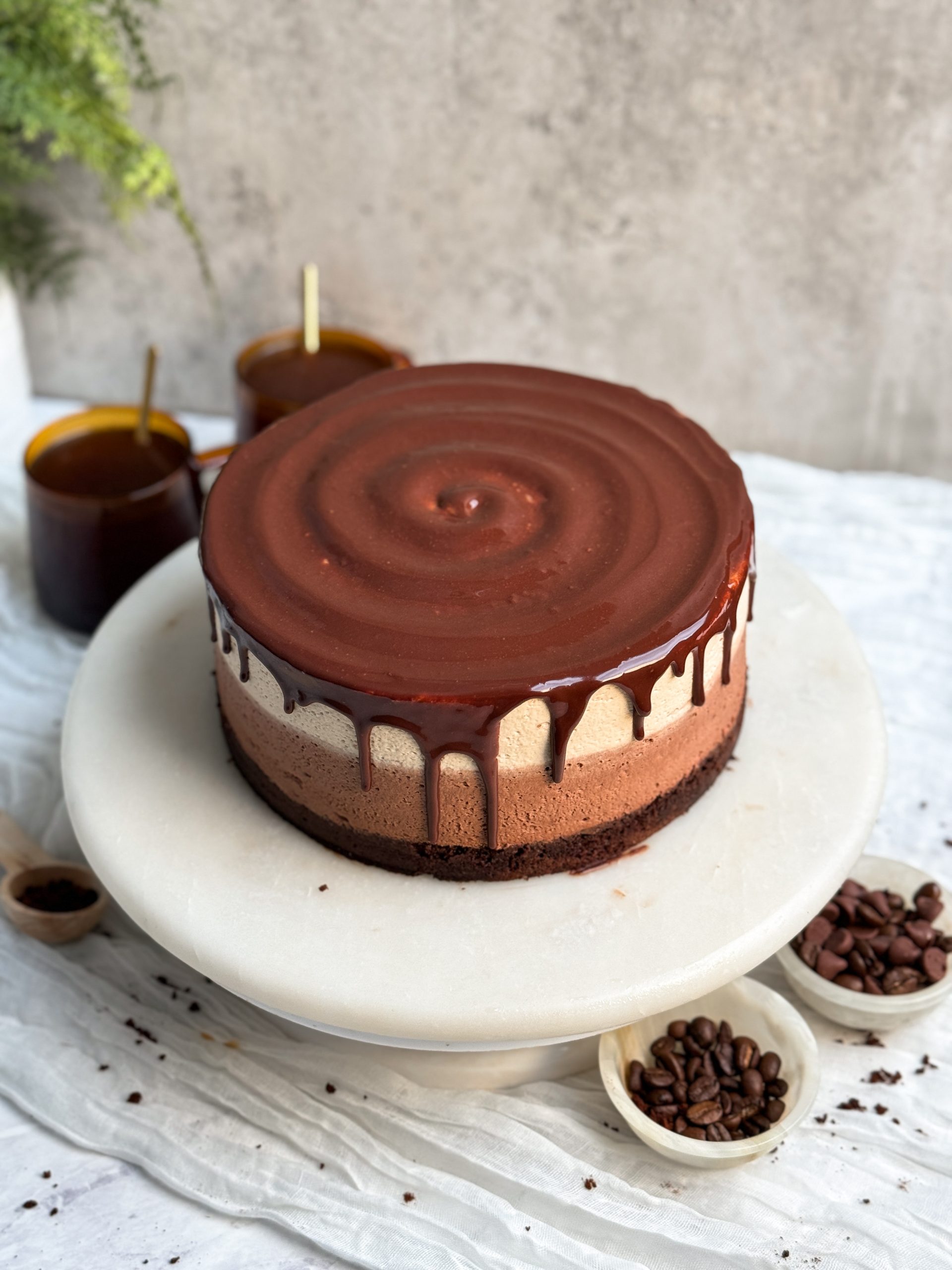 a layered mocha mousse cake on a marble cake stand. the cake has a layer of chocolate cake, a layer of chocolate mousse, a layer of white chocolate mocha mousse, and is topped off with a shiny chocolate glaze dripping on the sides