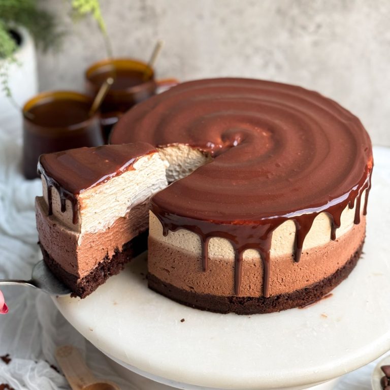a layered mocha mousse cake on a marble cake stand. the cake has a layer of chocolate cake, a layer of chocolate mousse, a layer of white chocolate mocha mousse, and is topped off with a shiny chocolate glaze dripping on the sides. A slice is being pulled out from the side