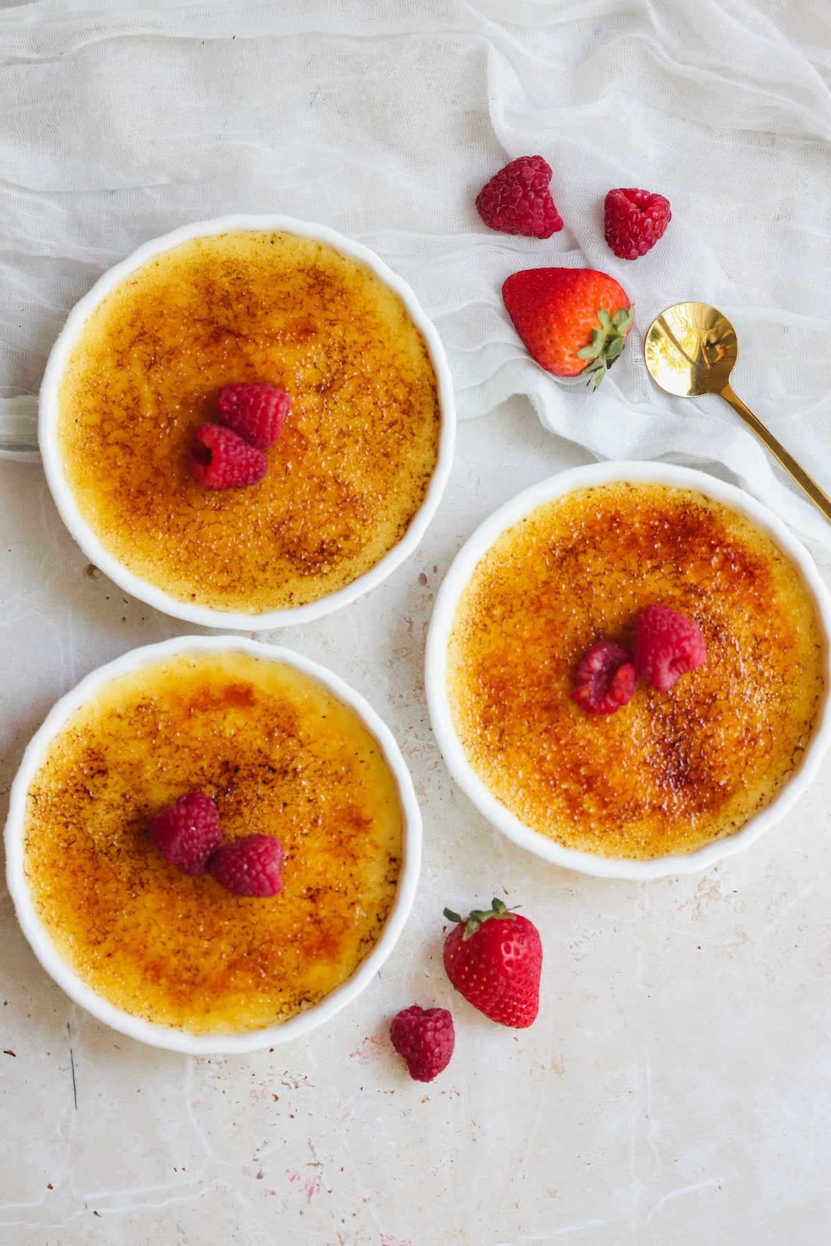 Three ramekins of this classic Creme Brulee recipe are topped with raspberries, surrounded by strawberries and raspberries, with a gold spoon and white cloth nearby on a light-colored surface.