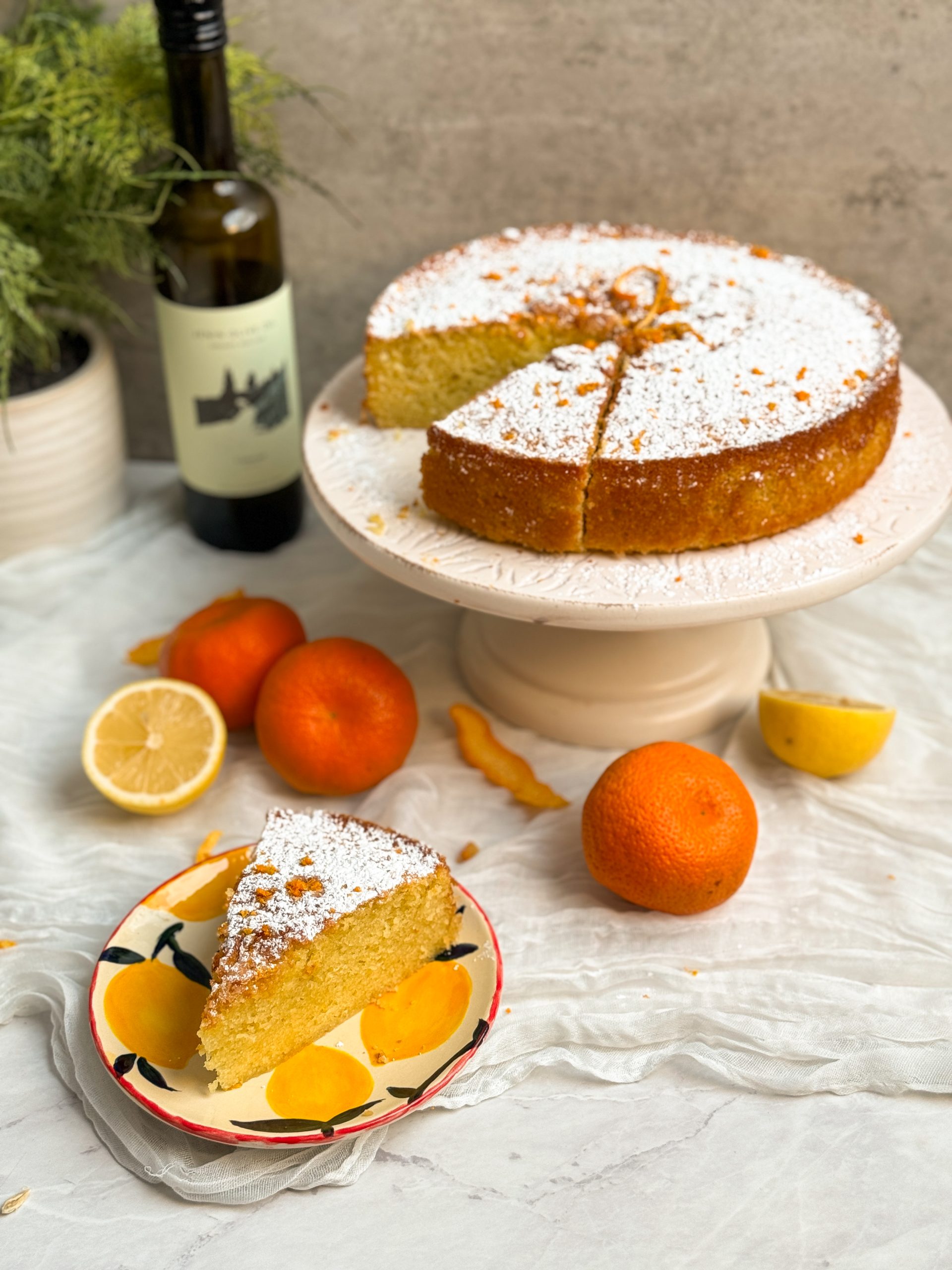orange olive oil cake on a white cake stand, dusted with powdered sugar and decorated with lemon zest. cake has a beautiful golden color. Oranges placed on the side, olive oil bottle in the back. Cake is sliced, and a slice is placed on a small plate in the front