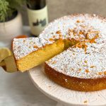 orange olive oil cake on a white cake stand, dusted with powdered sugar and decorated with lemon zest. cake has a beautiful golden color. Oranges placed on the side, olive oil bottle in the back. Cake is sliced and a spatula is pulling out a slice revealing the moist, tender interior