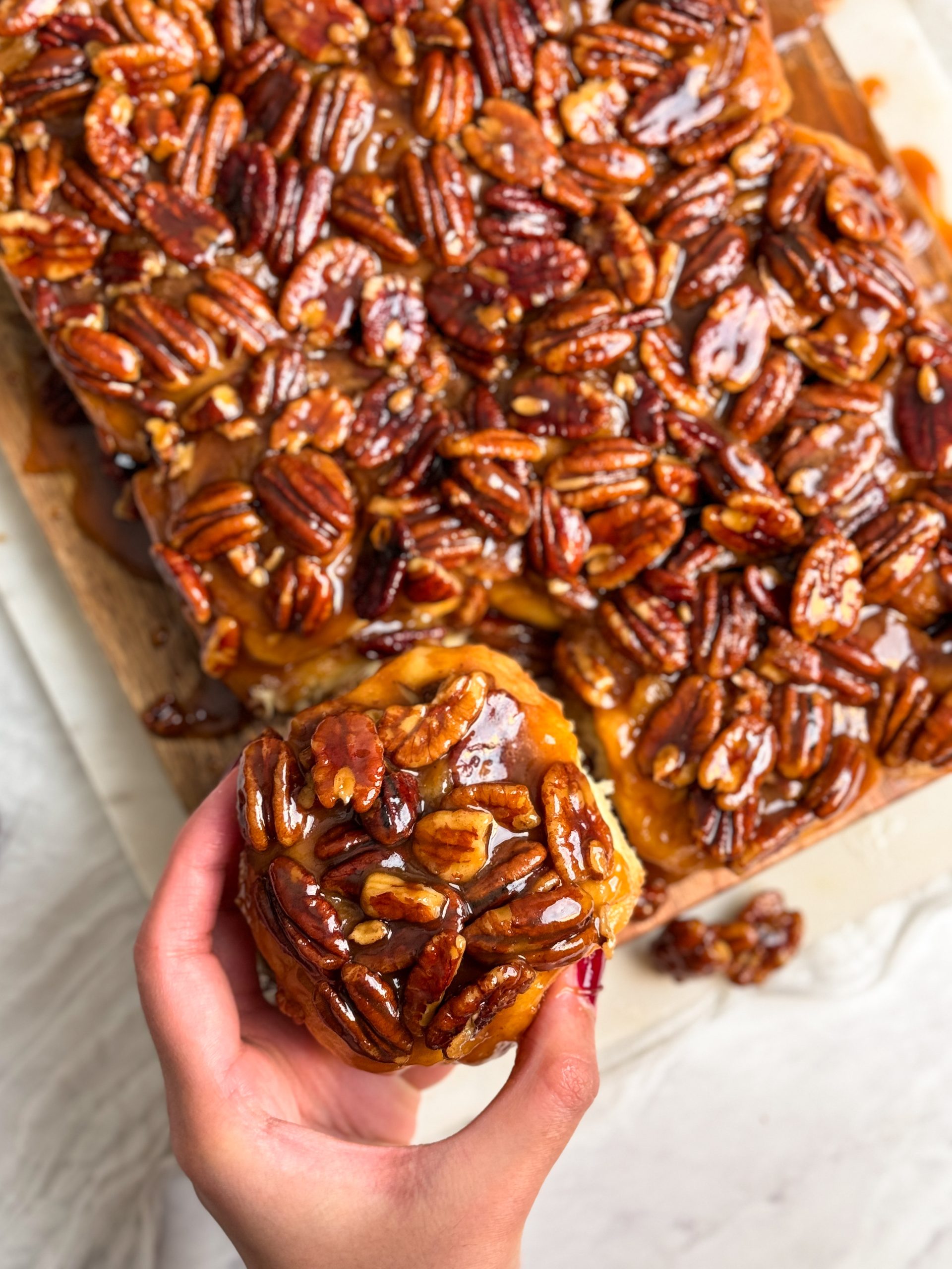 12 sticky pecan cinnamon buns with a caramel pecan topping on a wooden cutting board. they look soft and fluffy with a beautiful golden nutty topping. a hand is pulling out one bun which is in focus
