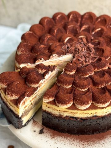 tiramisu cheesecake on a white marble cake stand. Cheesecake has a chocolate crust, a plain cheesecake filling, and is topped with dollops of mascarpone cream dusted with cocoa powder. A spatula is pulling out a slice, revealing the creamy interior and the layer of coffee soaked ladyfingers in the middle. Close up picture