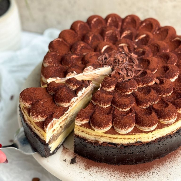 tiramisu cheesecake on a white marble cake stand. Cheesecake has a chocolate crust, a plain cheesecake filling, and is topped with dollops of mascarpone cream dusted with cocoa powder. A spatula is pulling out a slice, revealing the creamy interior and the layer of coffee soaked ladyfingers in the middle. Close up picture