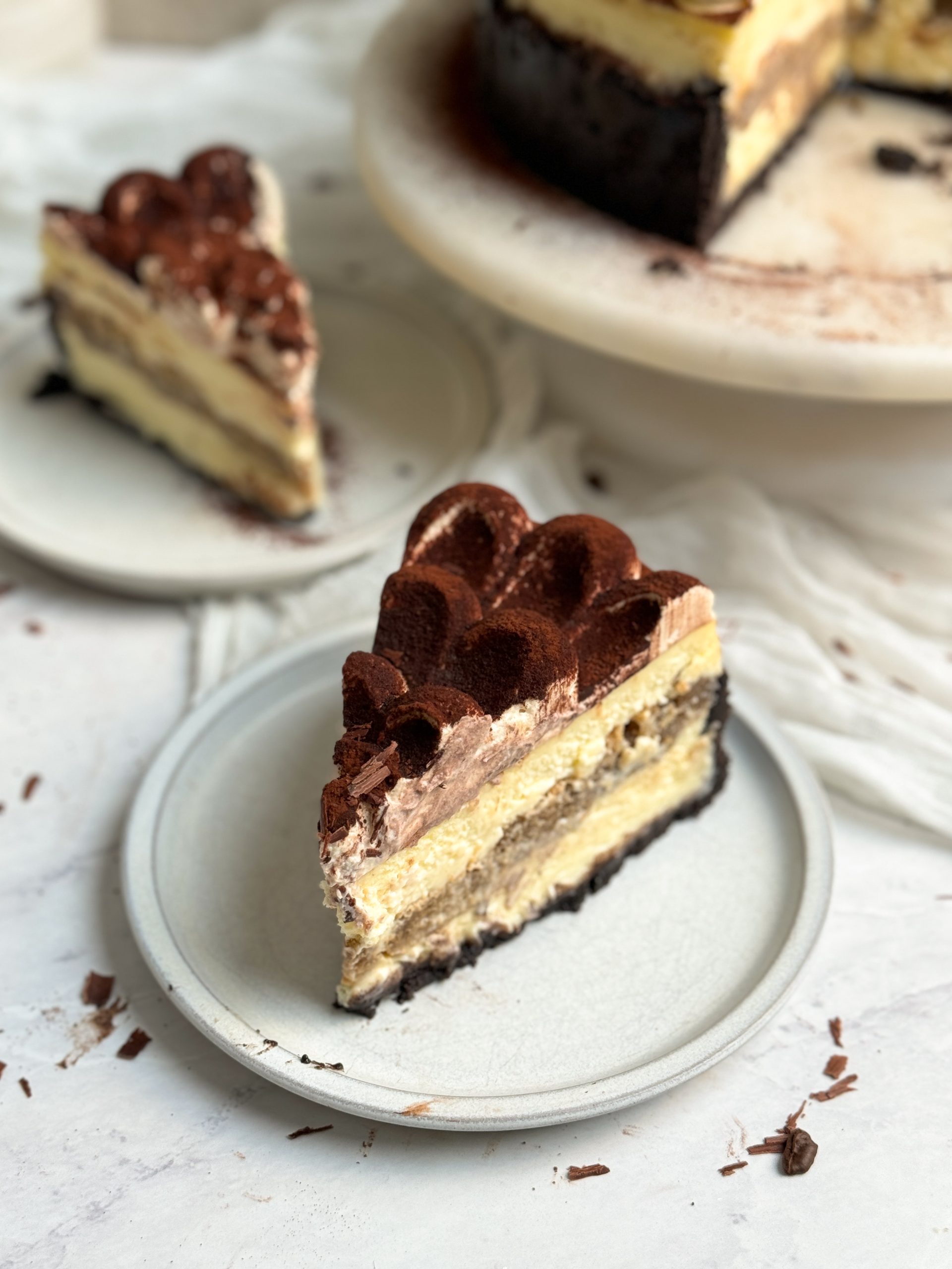 a slice of baked tiramisu cheesecake on a small plate, with a chocolate crust, creamy cheesecake interior, coffee soaked ladyfingers in the middle, and mascarpone cream with cocoa powder on top. Another slice in the background