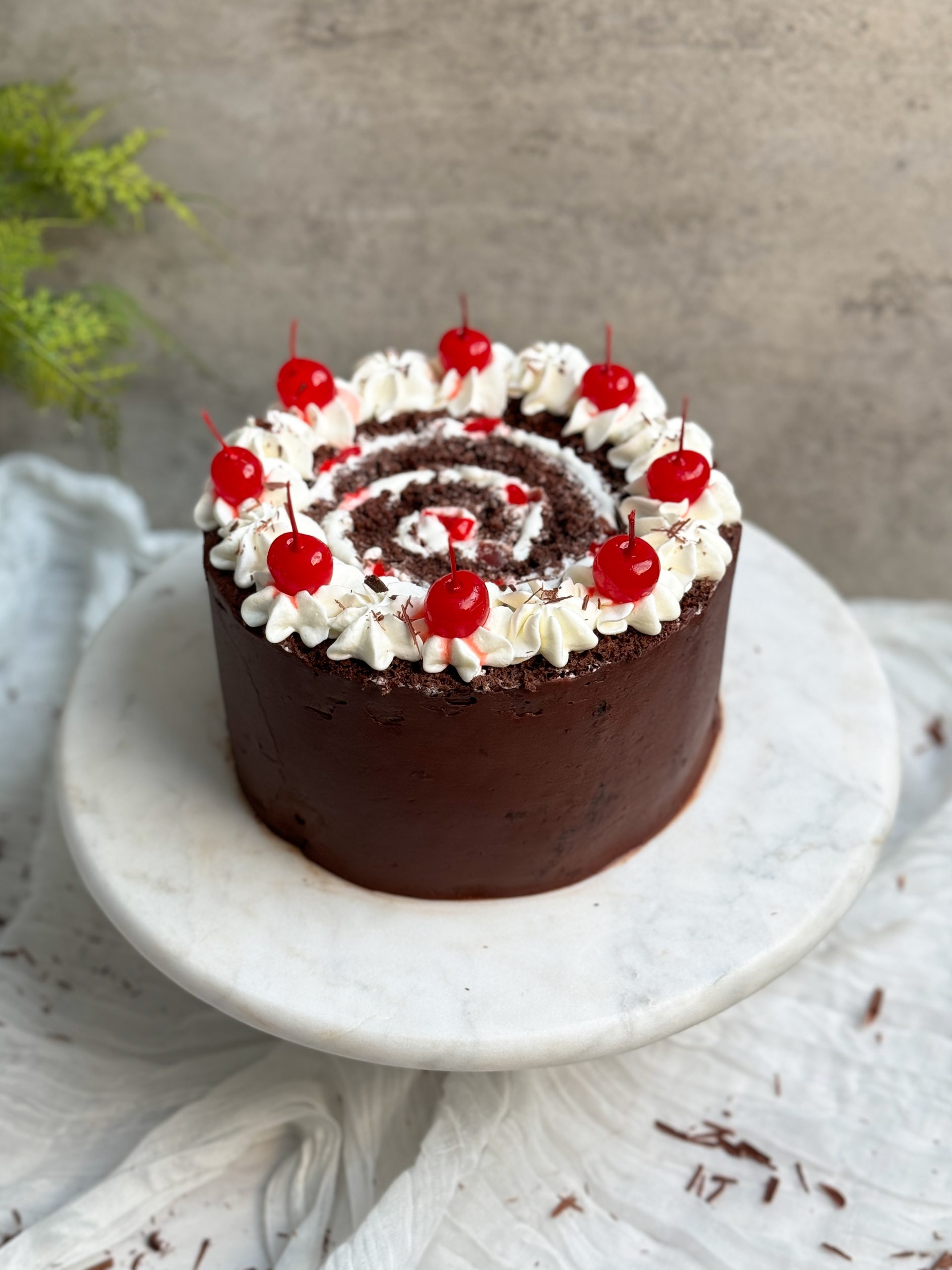 vertical black forest swiss roll cake on a marble serving stand. cake is coated in ganache with a swirl on top. decorated with whipped cream and cherries