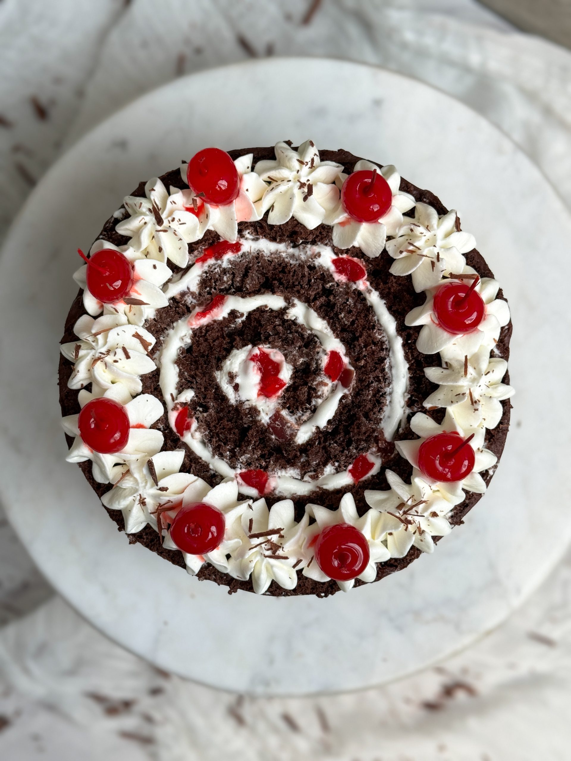 vertical black forest swiss roll cake from the top. the cake has a swirl of chocolate cake and whipped cream, with cream and cherries used for decoration on top