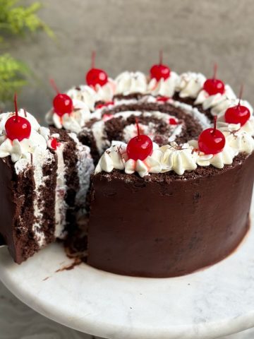 vertical black forest swiss roll cake on a marble serving stand. cake is coated in ganache with a swirl on top. a slice is being pulled out to reveal vertical layers of cake and whipped cream with cherries inside and on top