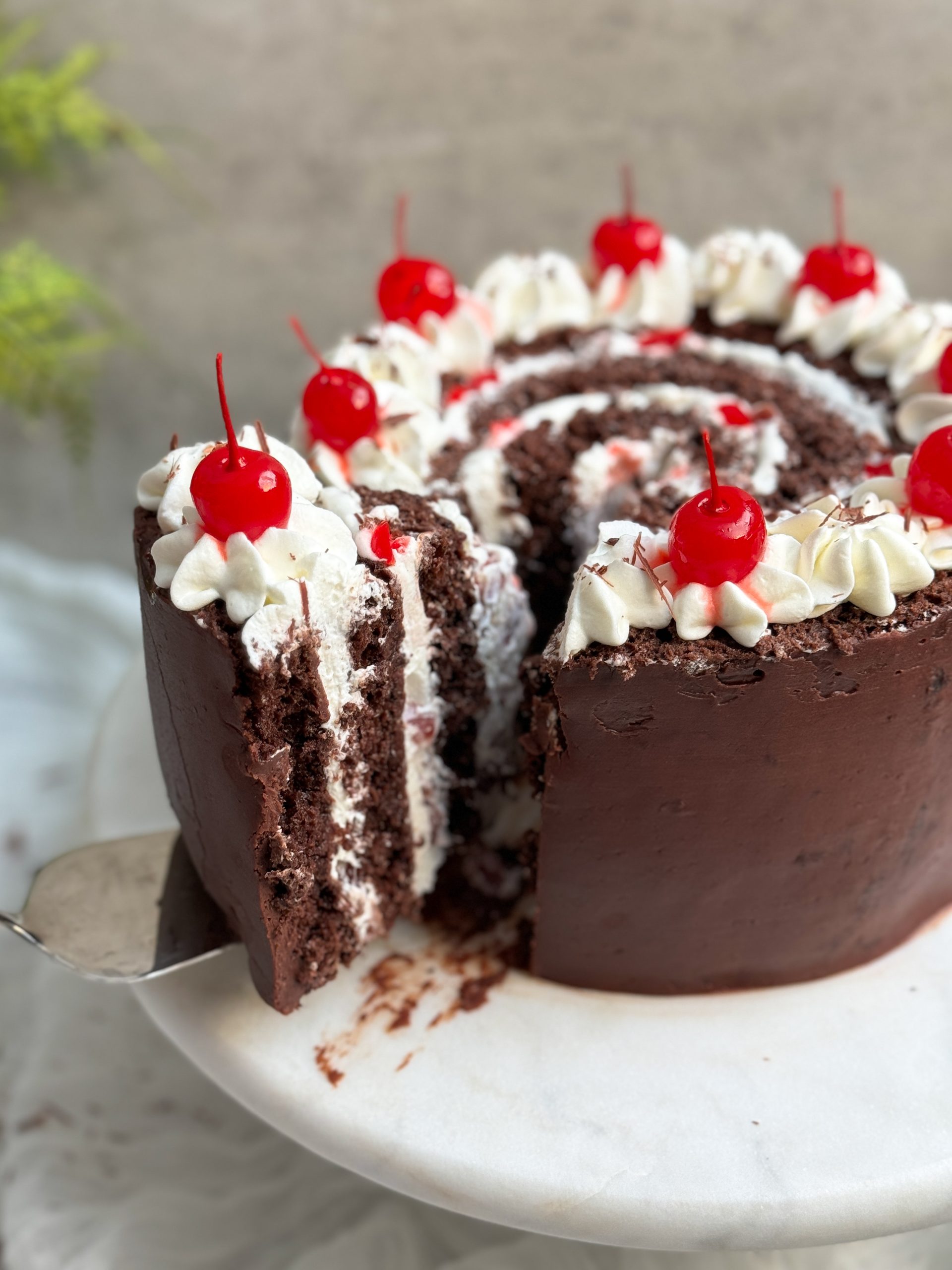 vertical black forest swiss roll cake on a marble serving stand. cake is coated in ganache with a swirl on top. a slice is being pulled out to reveal vertical layers of cake and whipped cream with cherries inside and on top. close up picture