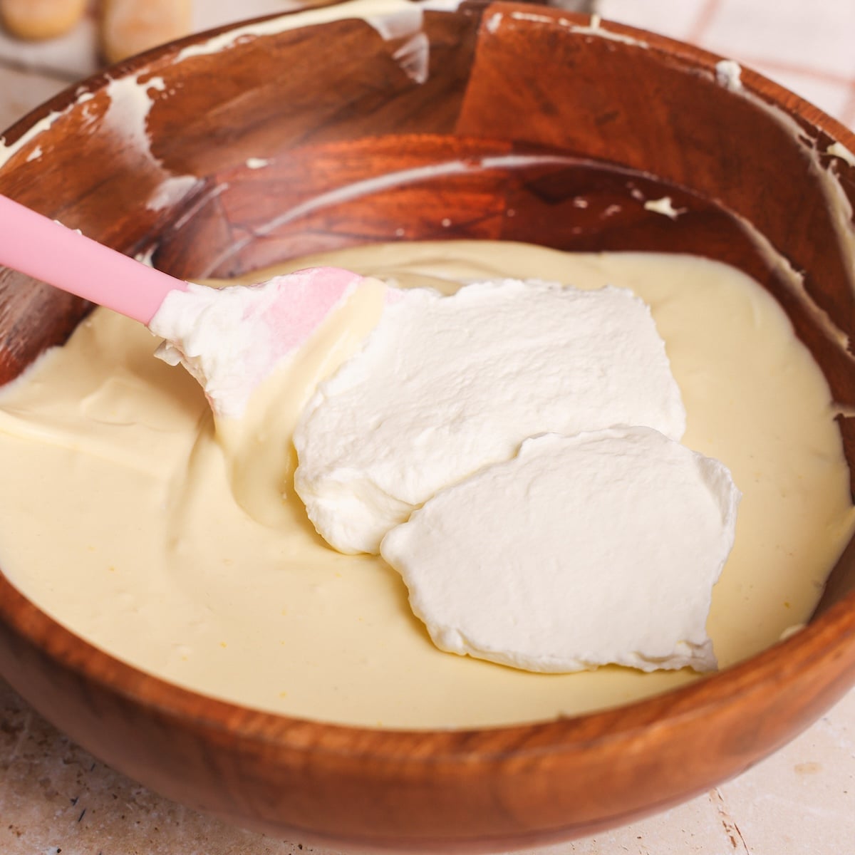 A pink spatula folds whipped cream into a bowl of yellow mixture in a wooden mixing bowl.