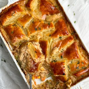 a rectangular dish with chicken pot pie inside. there are squares of golden crispy pastry arranged in a beautiful pattern on top of the pie, garnished with parsley. a portion has been taking out showing a creamy chicken and veggie filling