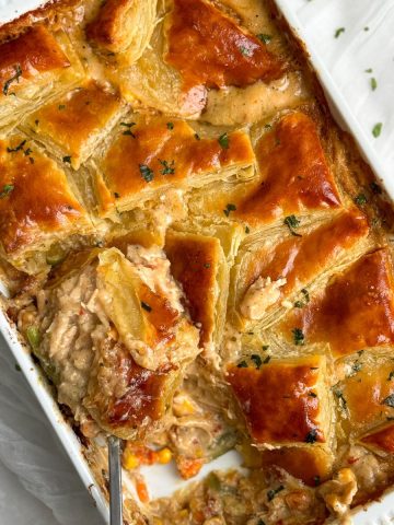 a rectangular dish with chicken pot pie inside. there are squares of golden crispy pastry arranged in a beautiful pattern on top of the pie, garnished with parsley. a portion has been taking out showing a creamy chicken and veggie filling