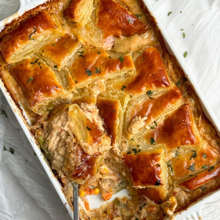 a rectangular dish with chicken pot pie inside. there are squares of golden crispy pastry arranged in a beautiful pattern on top of the pie, garnished with parsley. a portion has been taking out showing a creamy chicken and veggie filling