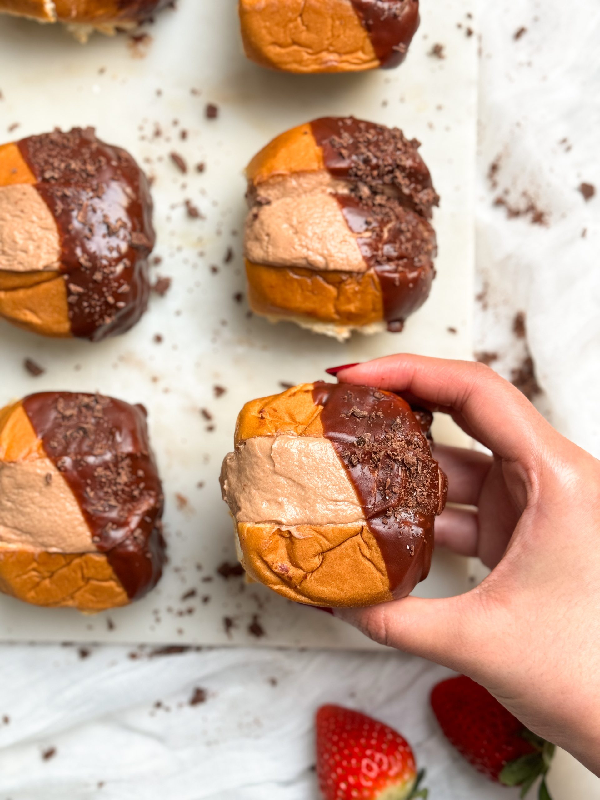 close up of chocolate cream buns on a marble serving board with a hand lifting one., the buns are dipped in a shiny chocolate glaze on the side
