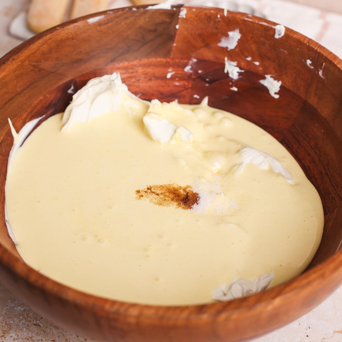 A wooden bowl filled with a creamy yellow egg yolk mixture, dollops of mascarpone cheese, and a sprinkle of vanilla in the center, sits on a light countertop