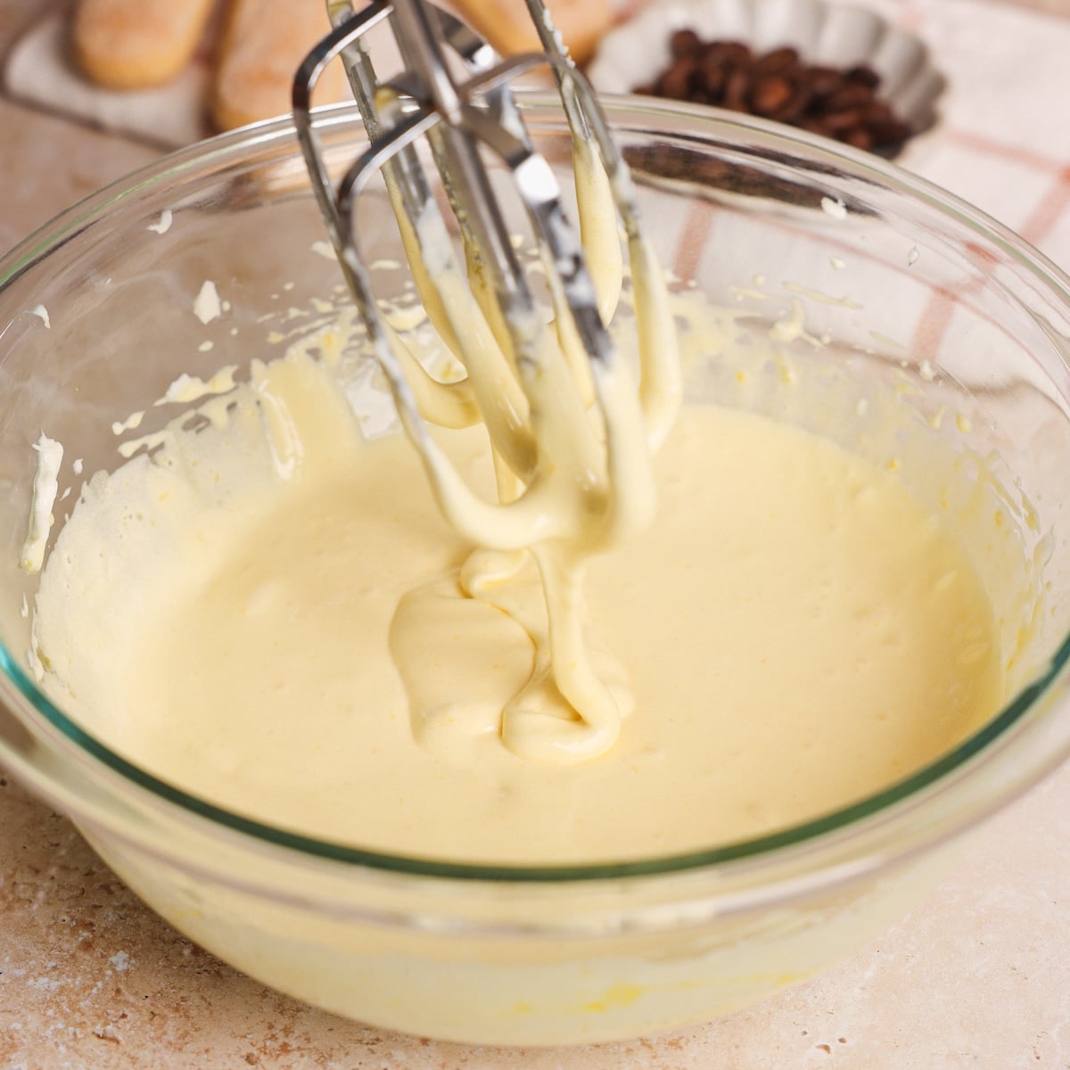 A glass bowl filled with creamy yellow egg yolks and sugar mixture is being mixed with electric beaters. Ingredients and a kitchen towel are visible in the background.