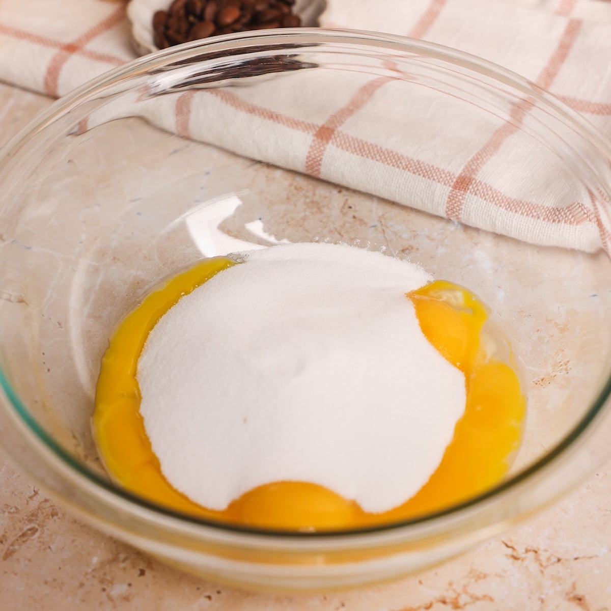 A glass bowl with egg yolks and a mound of white sugar inside, placed on a light countertop