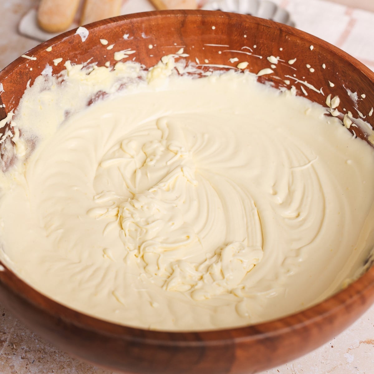 A wooden bowl filled with smooth, creamy yellow mascarpone and egg mixture sits on a countertop, with some batter splattered along the sides of the bowl