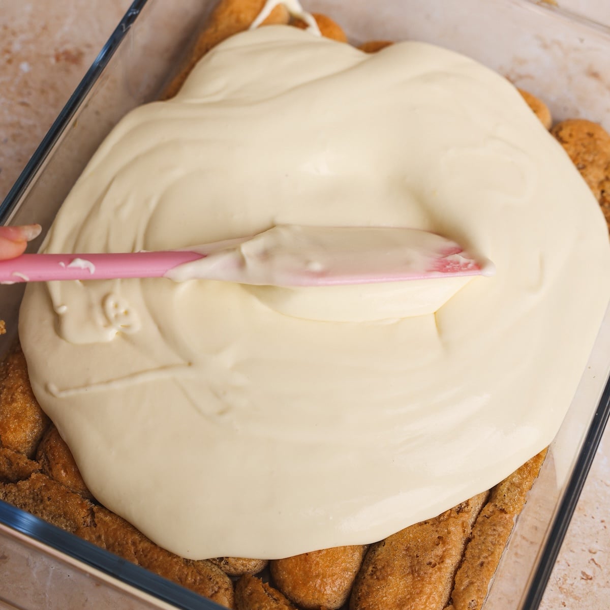 A hand uses a pink spatula to spread creamy mascarpone cream over a baked dessert in a glass dish, perfect for elevating your next tiramisu recipe.