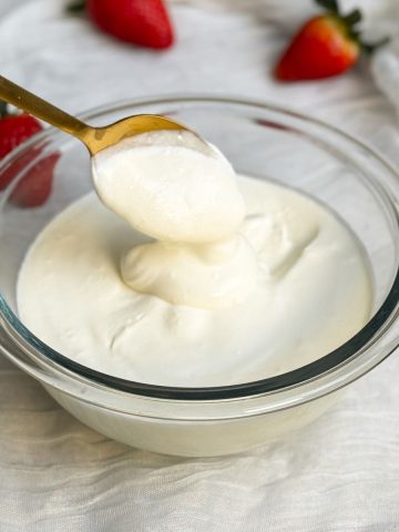 a glass bowl with homemade sour cream. sour cream is thick and creamy. a gold spoon is scooping up some sour cream to show how thick it is. sour cream is falling down in a dollop