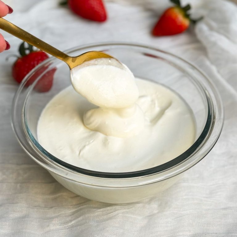 a glass bowl with homemade sour cream. sour cream is thick and creamy. a gold spoon is scooping up some sour cream to show how thick it is. sour cream is falling down in a dollop