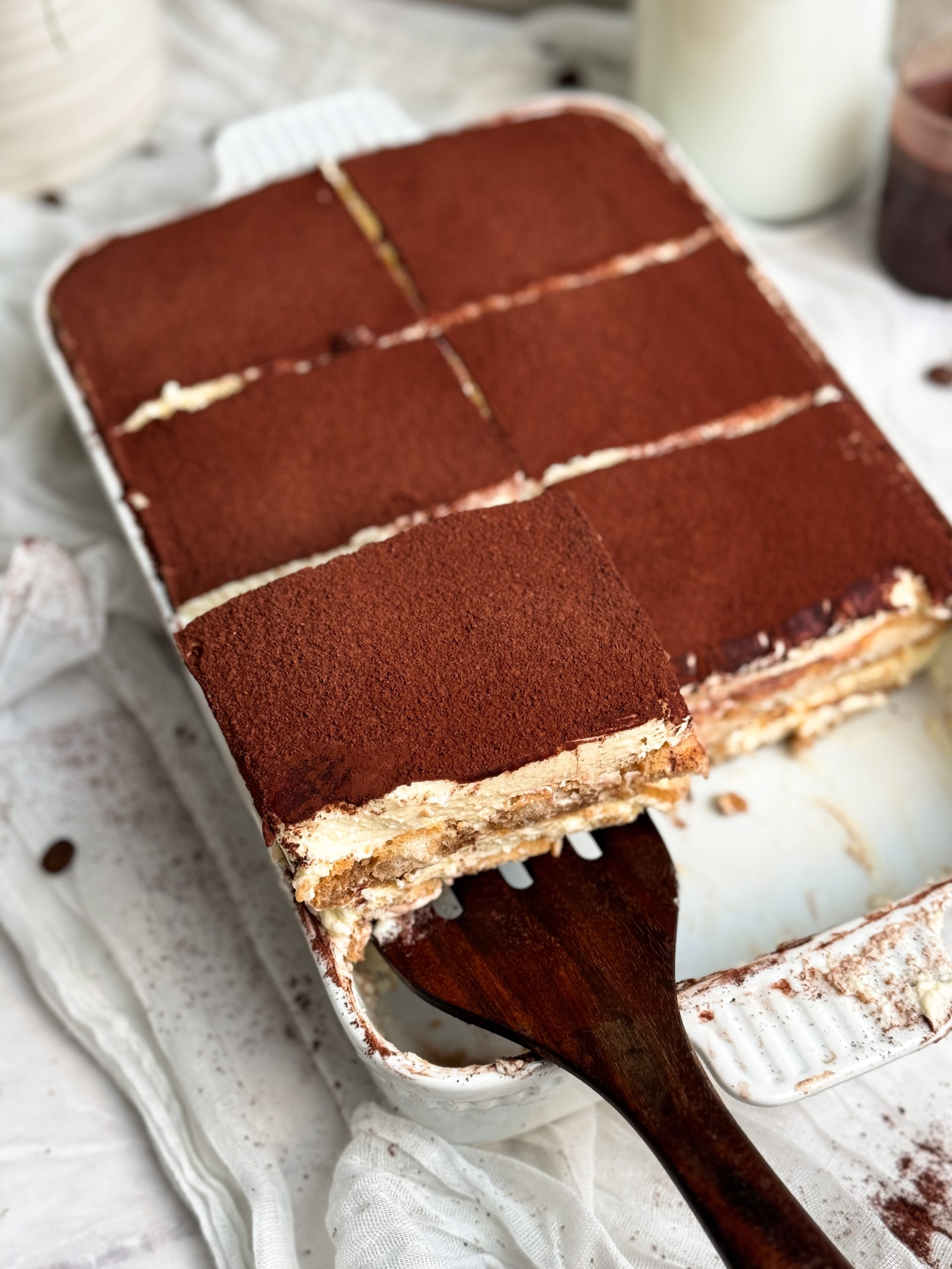 a spatula pulling out a slice of tiramisu from a pan, showing layers of cream and ladyfingers topped with cocoa powder. picture from the top