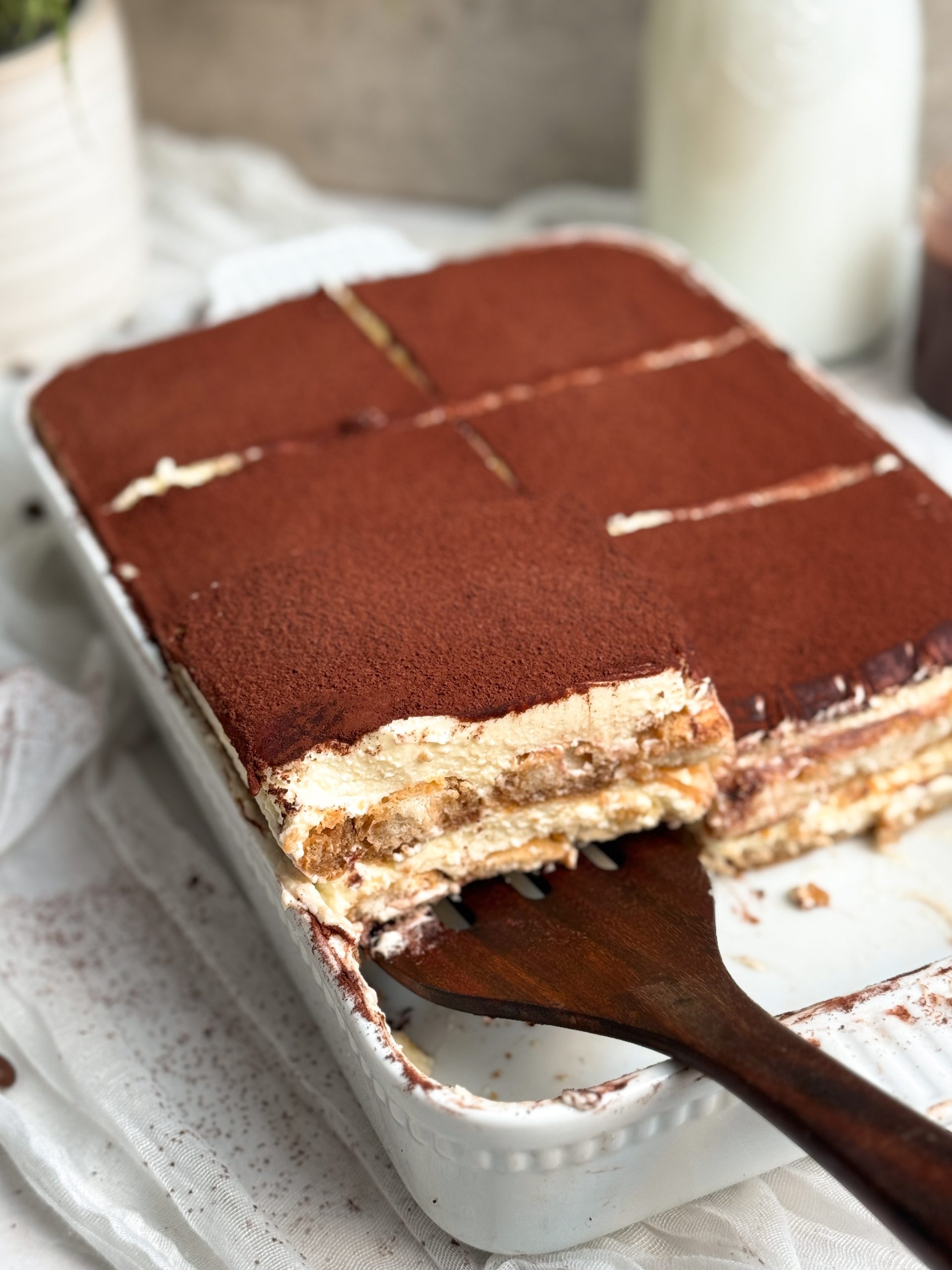 a spatula pulling out a slice of tiramisu from a pan, showing layers of cream and ladyfingers topped with cocoa powder
