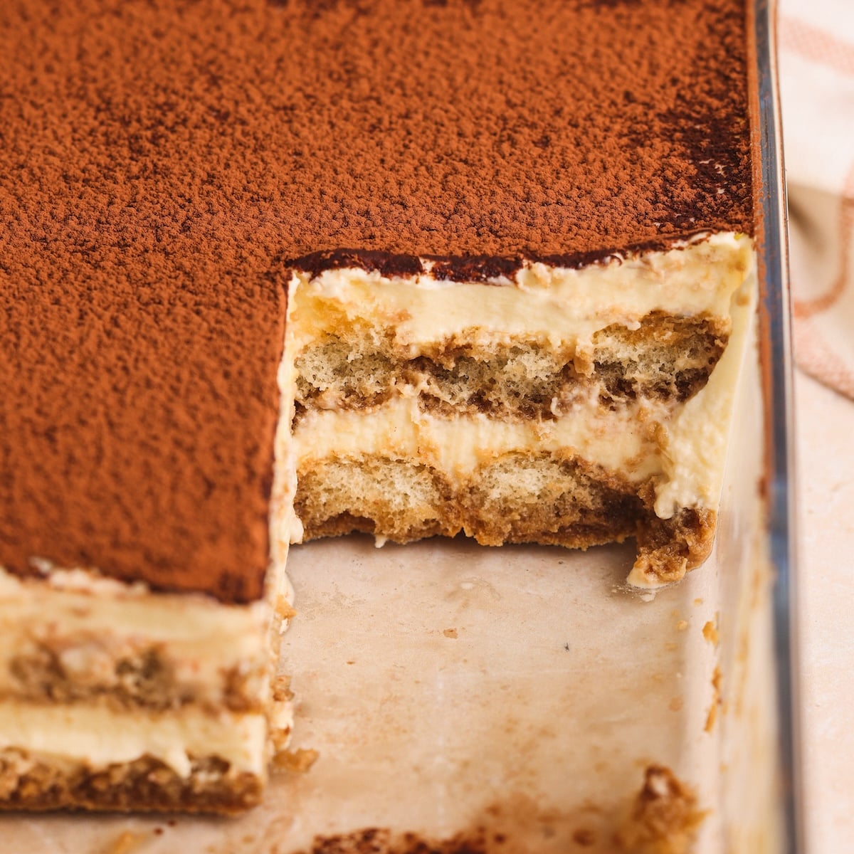 A close-up of a tiramisu dessert in a glass dish, with a square piece removed, showing layers of cream, coffee-soaked ladyfingers, and cocoa powder dusted on top. The layers are even and the cream looks smooth and creamy