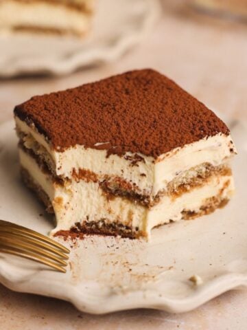 A square slice of tiramisu recipe topped with cocoa powder sits on a scalloped white plate, with a fork resting nearby and a bite taken out, revealing the dessert's creamy and layered textures.
