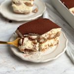 slice of tiramisu on a small plate with a bite being taken out, another slice in the back ground