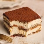 A close-up of a slice from a classic tiramisu recipe with two creamy layers and cocoa powder on top, showing a bite taken out of one corner. A gold fork rests nearby on a light plate.