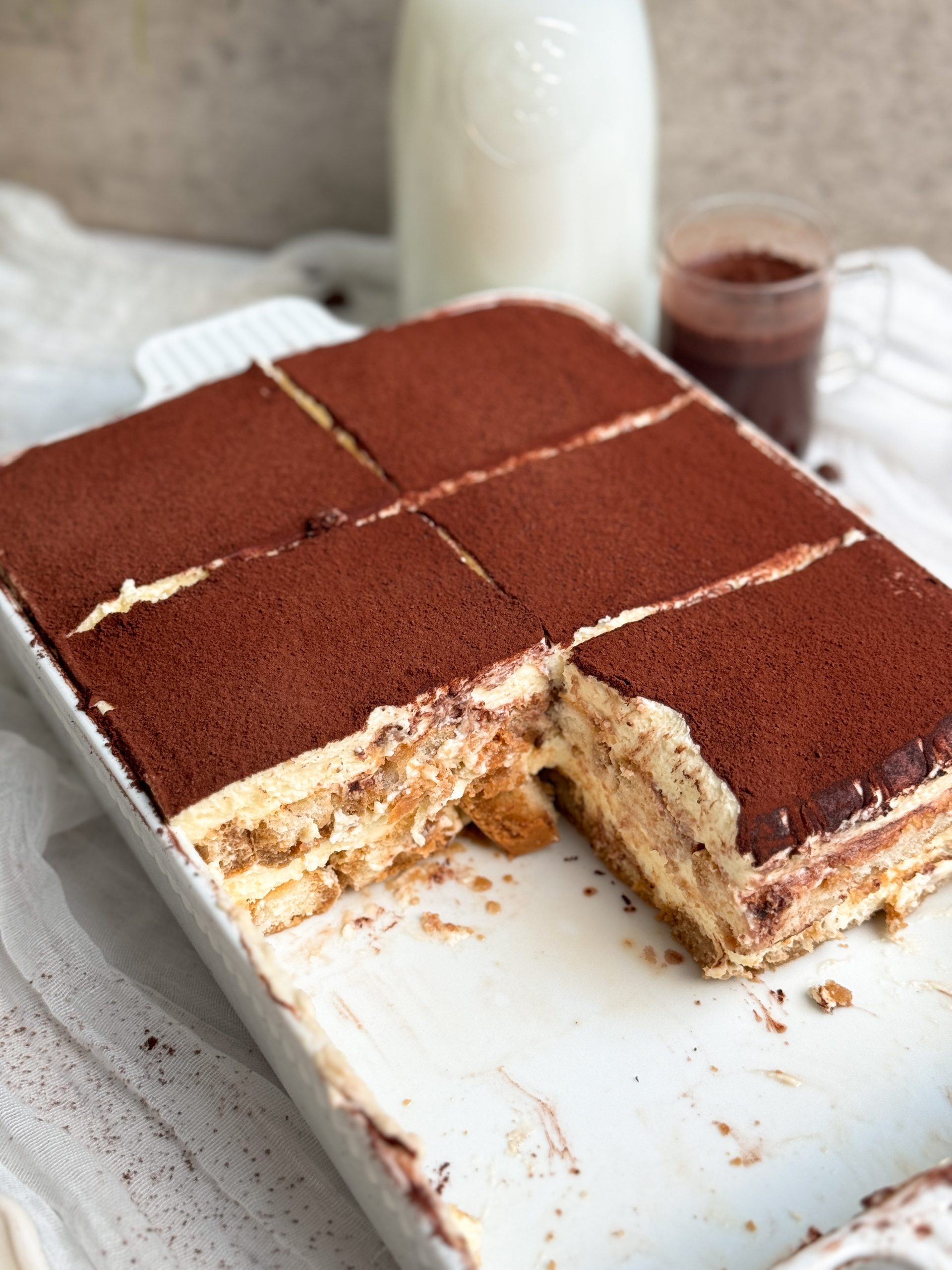 tiramisu in a white pan, cut into slices, with a slice taken out to show the layers of ladyfingers and mascarpone cream