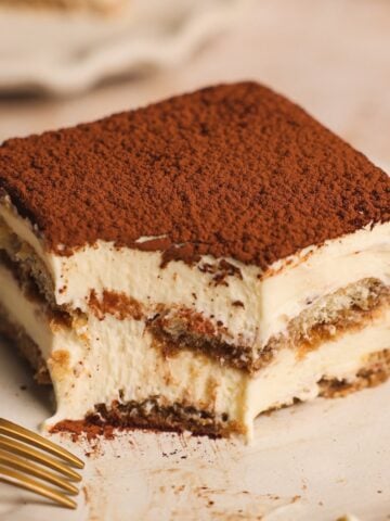 A close-up of a slice from a classic tiramisu recipe with two creamy layers and cocoa powder on top, showing a bite taken out of one corner. A gold fork rests nearby on a light plate.