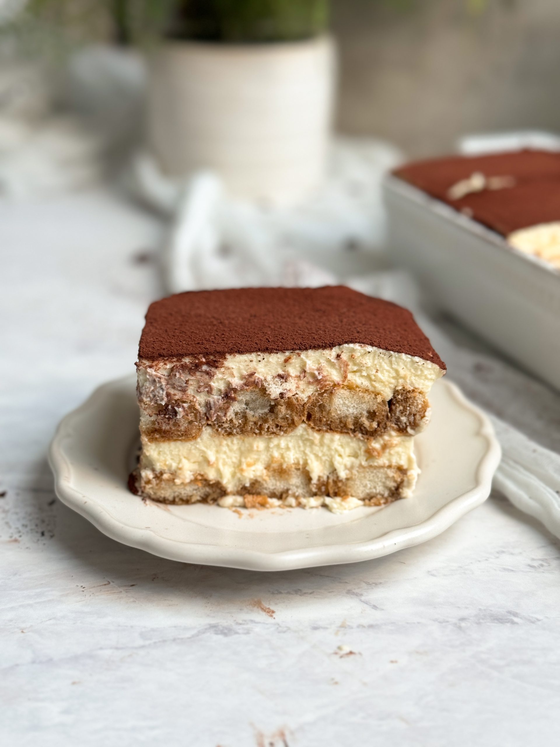 slice of tiramisu on a small plate, picture from the side to show layers of cream and ladyfingers, topped with cocoa powder