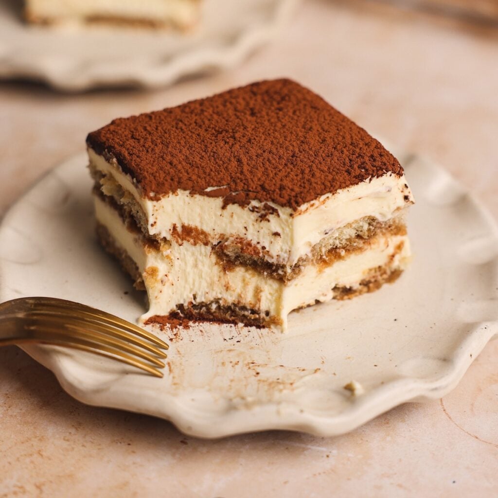 A partially eaten slice of tiramisu sits on a wavy white plate, topped with cocoa powder. Tiramisu looks very smooth and creamy, with a fork resting nearby.