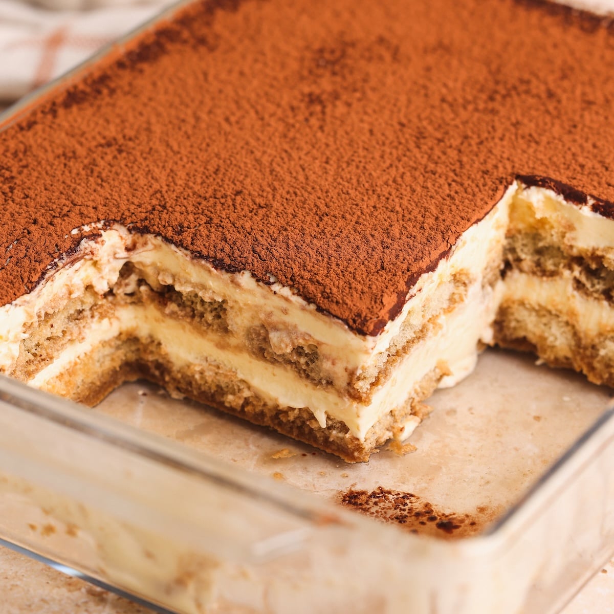 A glass dish of tiramisu with a missing corner, revealing layers of cream, coffee-soaked ladyfingers, and cocoa powder dusted on top