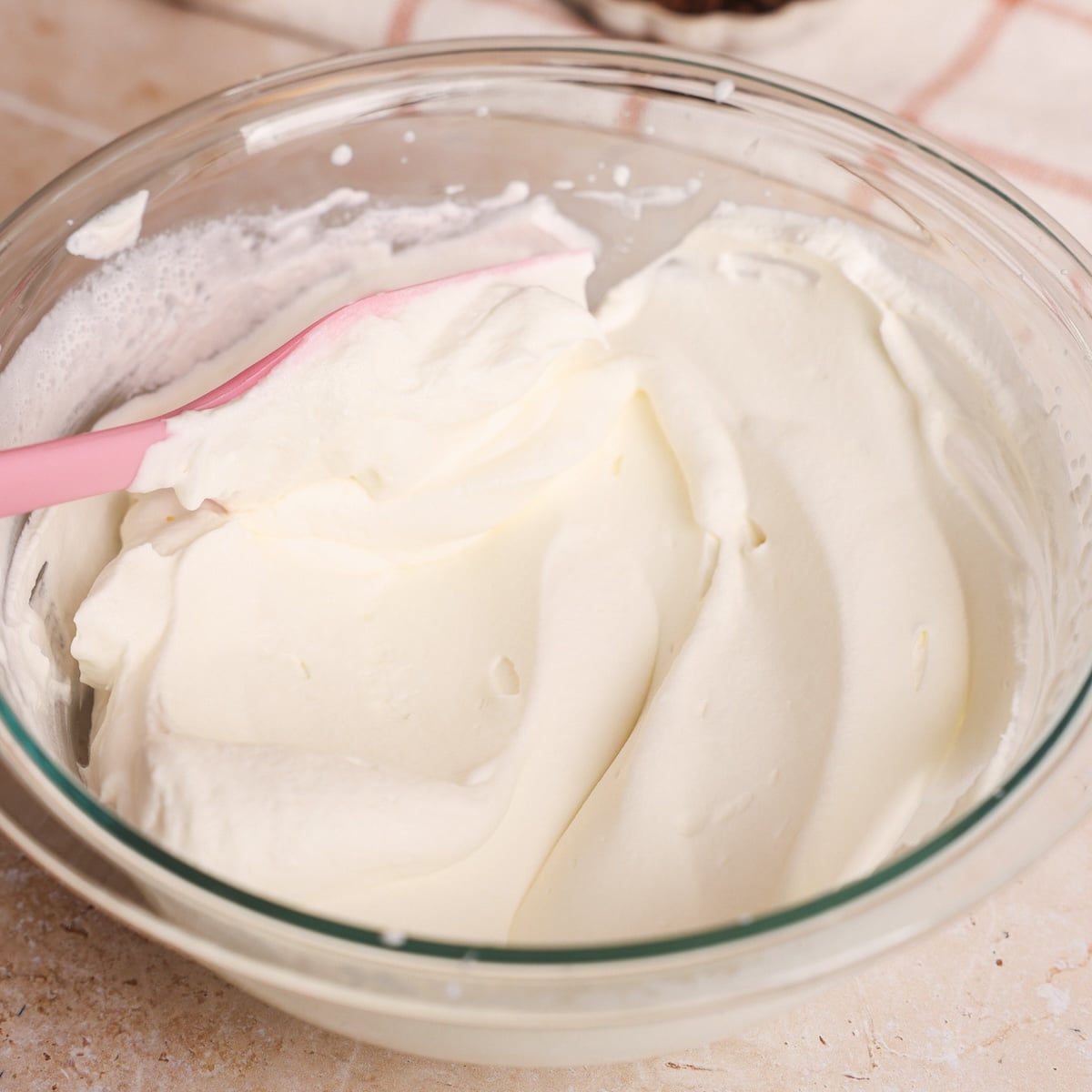 A glass bowl filled with smooth, whipped crea and a pink spatula resting inside, on a light-colored countertop.