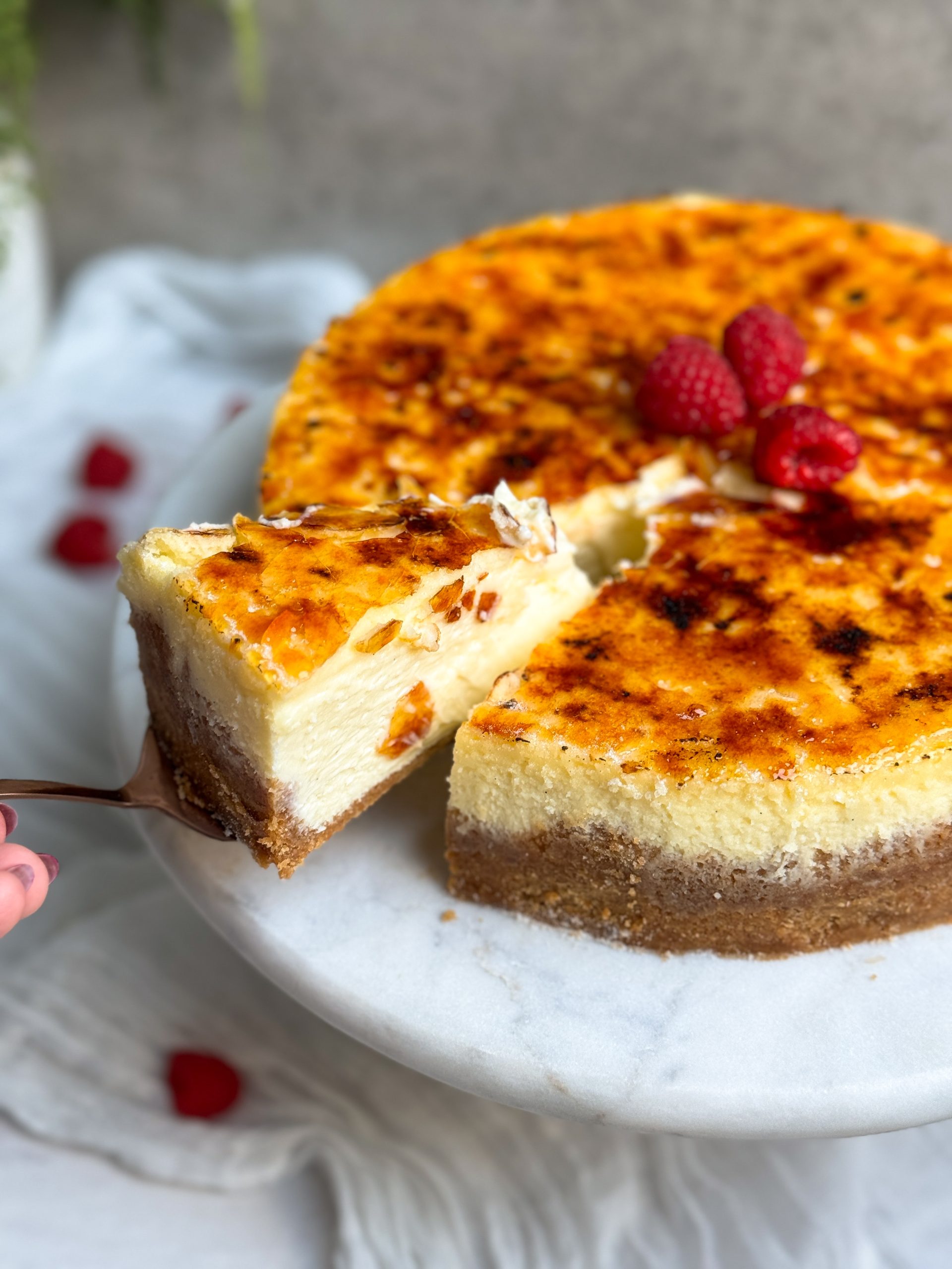 creme brulee cheesecake with a golden crispy top on a marble serving board, decorated with some raspberries. a spatula is pulling out a slice showing the creamy texture of the cheesecake