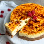 creme brulee cheesecake with a golden crispy top on a marble serving board, decorated with some raspberries. a spatula is pulling out a slice showing the creamy texture of the cheesecake; picture from a higher angle