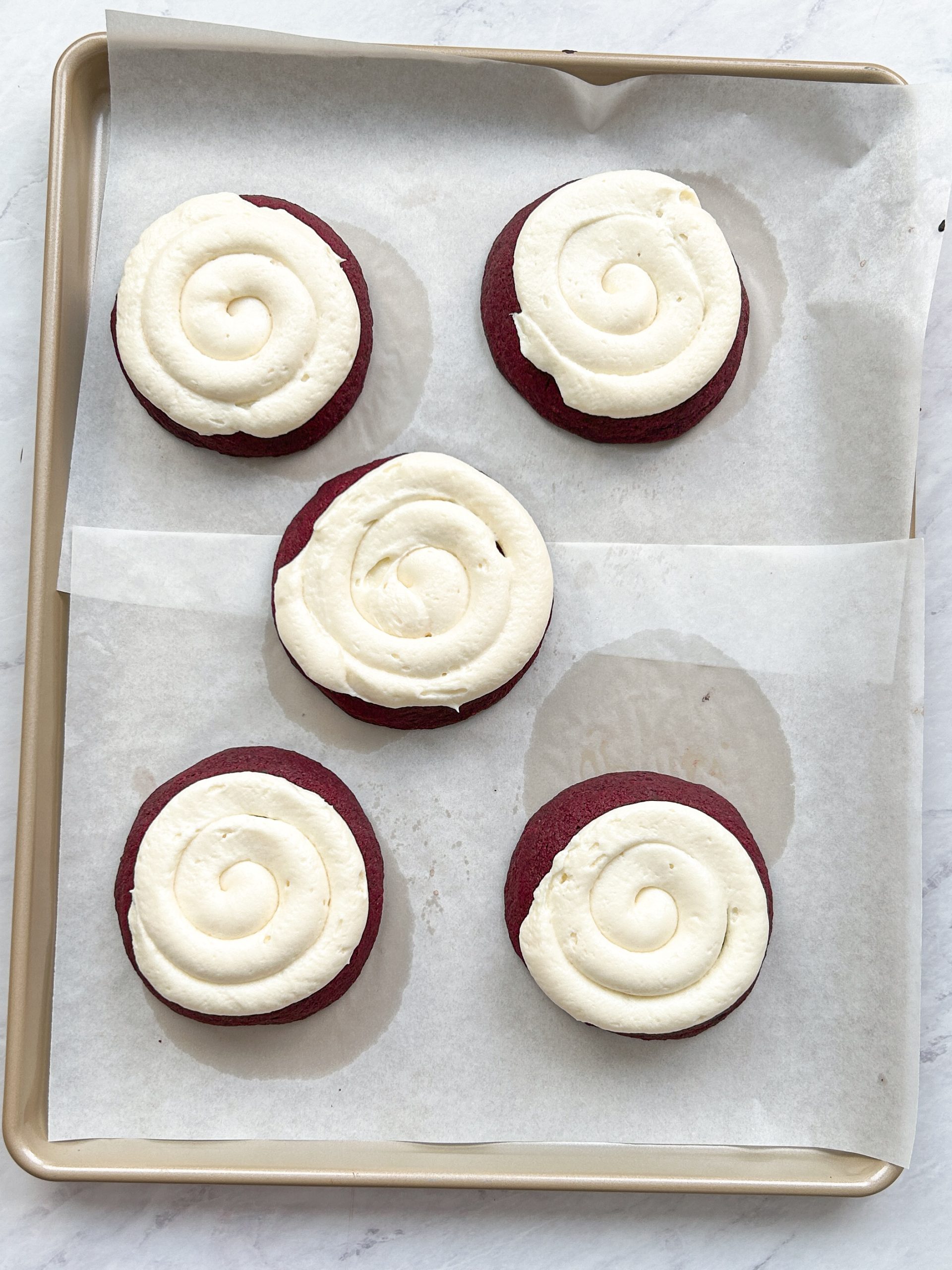 5 red velvet cookies with cream cheese frosting swirled on top on a cookie sheet