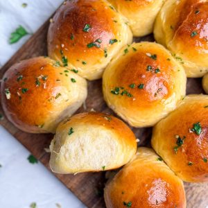 close up of garlic butter dinner rolls on a wooden serving board. Rolls have a beautiful golden shine on them and have been brushed generously with garlic butter. one roll is placed sideways showing the soft interior texture