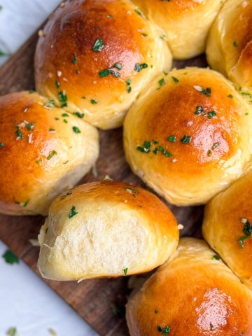 close up of garlic butter dinner rolls on a wooden serving board. Rolls have a beautiful golden shine on them and have been brushed generously with garlic butter. one roll is placed sideways showing the soft interior texture