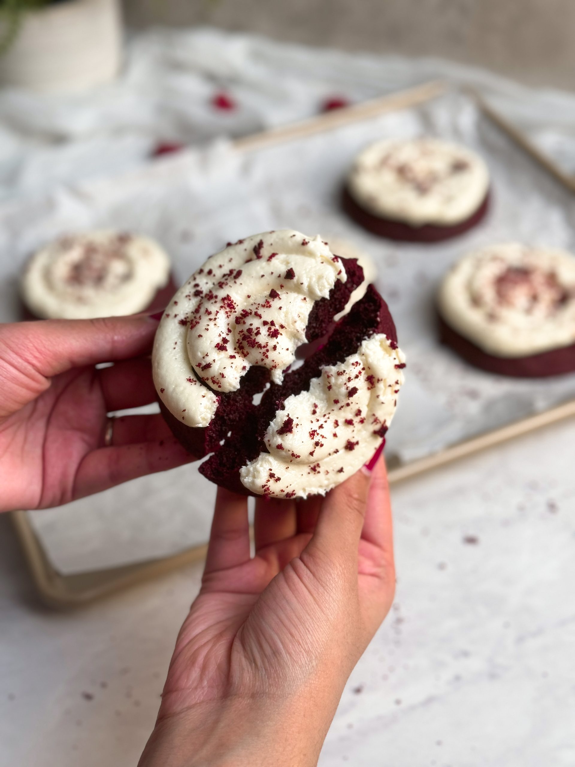 hand holding red velvet cookie with cream cheese frosting on top, cutting it into half to show the soft texture