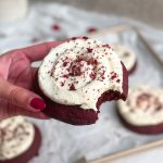 hand holding red velvet cookie with cream cheese frosting on top with a bite taken out of it